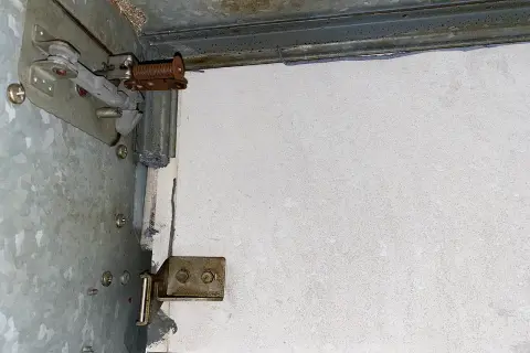 Detailaufnahme einer Brandschutzklappe mit sichtbaren mechanischen Teilen und einer weißen Platte. Möglicherweise Asbest-verdächtige Materialien.