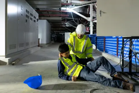 Ein Arbeiter hilft einem Kollegen, der nach einem Stromschlag auf dem Boden liegt. Der helfende Arbeiter trägt eine Warnweste und einen Helm.