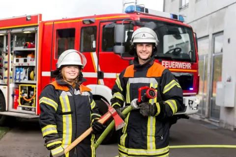 Zwei Feuerwehrleute vor einem roten Feuerwehrauto. Sie tragen Schutzkleidung und Helme. Die Frau hält eine Axt, der Mann einen Schlauch. So wird man Feuerwehrmann oder Feuerwehrfrau.