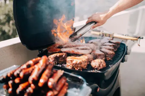 Leckeres Grillen auf dem Balkon: Würstchen und Steaks brutzeln auf dem Rost, Flammen lodern.