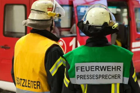 Zwei Feuerwehrleute stehen vor einem roten Fahrzeug. Einer trägt eine gelbe Weste mit der Aufschrift "Einsatzleiter", der andere eine grüne Weste mit der Aufschrift "FEUERWEHR PRESSESPRECHER".