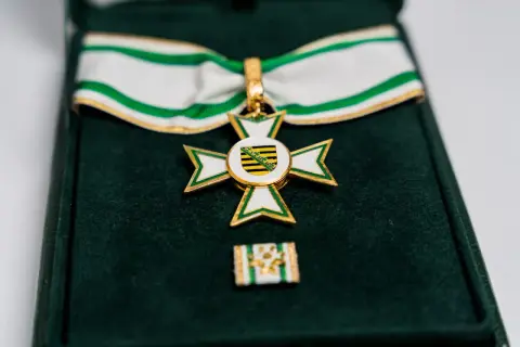 Ein goldenes Kreuz mit weißer und grüner Emaille, das ein Wappen mit schwarzen und gelben Streifen zeigt. Dieses Feuerwehr Ehrenzeichen liegt in einer grünen Samtschachtel.