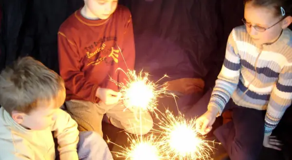 Kinder halten Wunderkerzen. Ein Junge im roten Pullover, ein Mädchen mit Brille und ein kleiner Junge im hellen Pulli genießen den Zauber. Ein schöner Moment, der an die Grundschule erinnert.