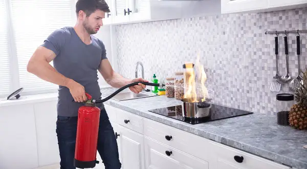 Mann löscht Topfbrand in der Küche mit Feuerlöscher. Dies ist ein Beispiel für Brandklasse F, Fettbrand.