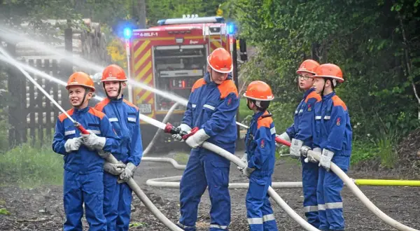 Jugendfeuerwehr übt praktische Ausbildungsideen: Kinder in blauen Uniformen und Helmen halten Wasserschläuche, ein Feuerwehrauto im Hintergrund.