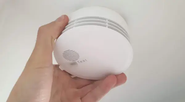Ein weißer Rauchmelder wird im Keller von einer Hand gehalten. Er hat einen Testknopf und Lüftungsschlitze.