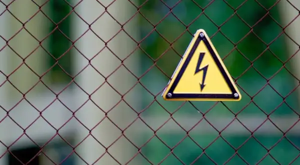 Gefahr! Gelbes Warnschild mit Blitzsymbol auf Drahtzaun. Bei einem leichten Stromschlag was tun? Sofort Abstand halten und Hilfe holen.