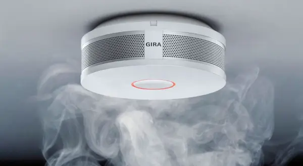 Ein Gira Rauchmelder mit orange blinkendem Ring, umgeben von aufsteigendem Rauch.