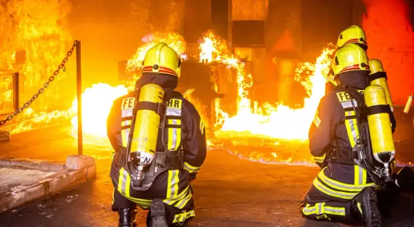 Zwei Feuerwehrleute in voller Montur knien vor einem intensiven Feuer. Ihre Ausbildung bei der Berufsfeuerwehr bereitet sie auf solche Einsätze vor.