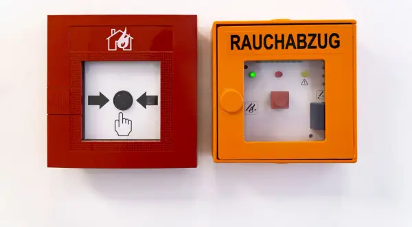 RWA Anlagen Wartung: Rote Brandmeldeanlage und orange Rauchabzugseinheit an weißer Wand.