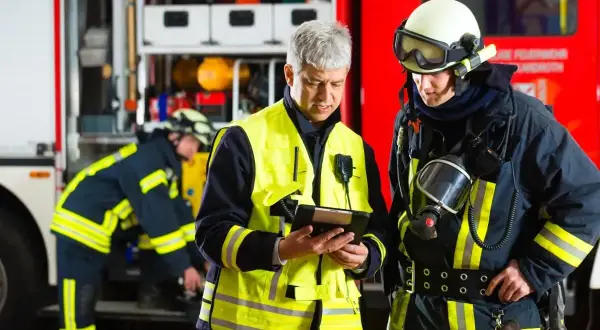 Feuerwehrleute besprechen Einsatzdetails. Ein Mann in Warnweste prüft ein Tablet, während ein Kollege in voller Montur mit Atemschutzmaske daneben steht. UVV Feuerwehr wird beachtet.