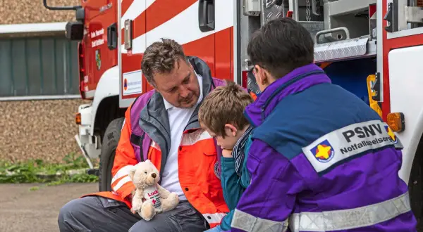 Ein Mann mit einem Teddybär spricht mit einem Kind neben einem Feuerwehrauto. Eine Person mit PSNV-Aufschrift auf der Jacke sitzt daneben.