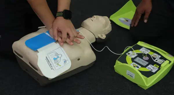 Schulung an einem Trainings-Defibrillator mit Reanimationspuppe. Die Hände üben die Herzdruckmassage. Regelmäßige **Defibrillator Wartung** ist wichtig.
