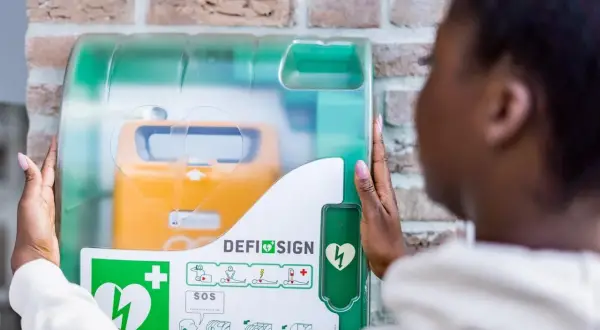 Frau öffnet Defibrillator-Gehäuse, um den Standort zu finden. Gelbes Gerät mit Herzsymbol sichtbar.