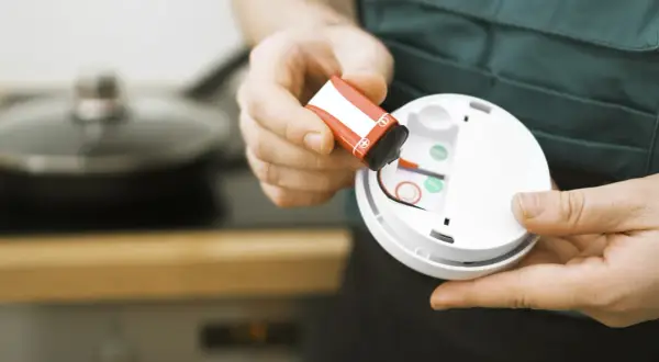 Batteriewechsel am Rauchmelder: Eine Hand hält eine rote 9V-Batterie, bereit zum Einsetzen in das weiße Gerät.