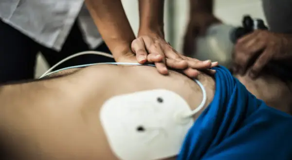 Hände führen eine Herzmassage durch, während ein AED-Einsatz vorbereitet wird. Ein weißes Elektrodenthermometer ist auf der Brust platziert.