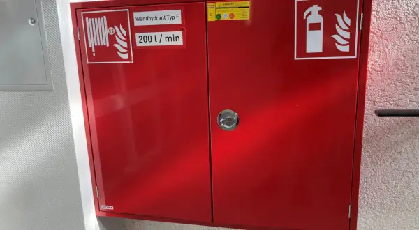 Roter Schrank für Brandschutz in einer Arztpraxis: Wandhydrant Typ F, 200 l/min, und Feuerlöscher-Symbol.