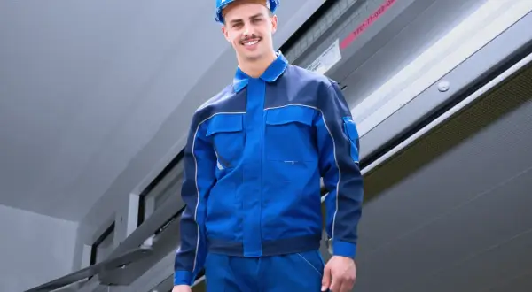 Ein Handwerker in blauer Arbeitskleidung und Helm trägt einen Werkzeugkasten. Er ist bereit für den Einsatz nach DIN 14677.