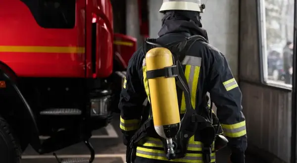 Ein Feuerwehrmann in voller Montur mit Atemschutzgerät steht bereit bei der Feuerwehr. Links im Bild ist ein Teil eines roten Feuerwehrautos zu sehen.