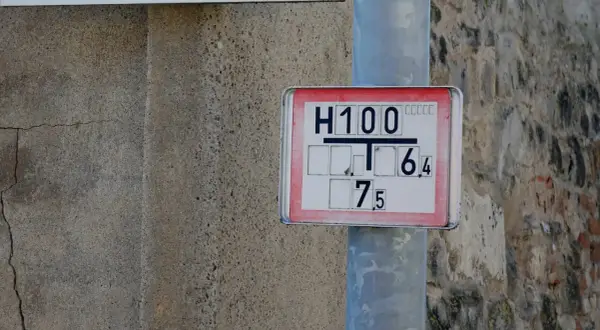 Nahaufnahme eines Hydrantenschilds mit der Aufschrift "H100 T 6,4 7,5". Das Schild ist an einem Metallpfosten befestigt, daneben eine raue Betonwand.