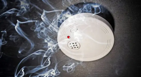 Ein weißer rauchmelder oder feuermelder mit roter LED auf dunklem Grund, umgeben von aufsteigendem Rauch.