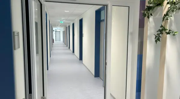 Langer Flur mit vielen Türen, beleuchtet von Deckenlampen. Grüne Notausgangsschilder sind sichtbar. Die Wartung von Feststellanlagen ist hier essenziell für die Sicherheit.