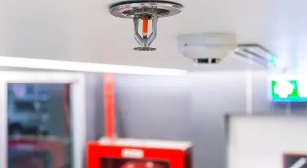 Eine Sprinkleranlage an der Decke, bereit für den Notfall. Daneben ein Rauchmelder und ein grünes Notausgangsschild.