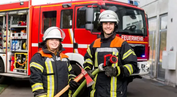 Zwei Feuerwehrleute vor einem roten Feuerwehrauto. Sie tragen Schutzkleidung und Helme. Die Frau hält eine Axt, der Mann einen Schlauch. So wird man Feuerwehrmann oder Feuerwehrfrau.