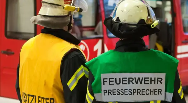 Zwei Feuerwehrleute stehen vor einem roten Fahrzeug. Einer trägt eine gelbe Weste mit der Aufschrift "Einsatzleiter", der andere eine grüne Weste mit der Aufschrift "FEUERWEHR PRESSESPRECHER".