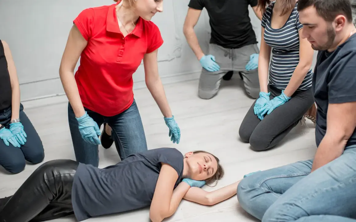 Erste-Hilfe-Kurs: Eine Frau wird in die stabile Seitenlage gebracht. Mehrere Personen mit blauen Handschuhen beobachten aufmerksam.