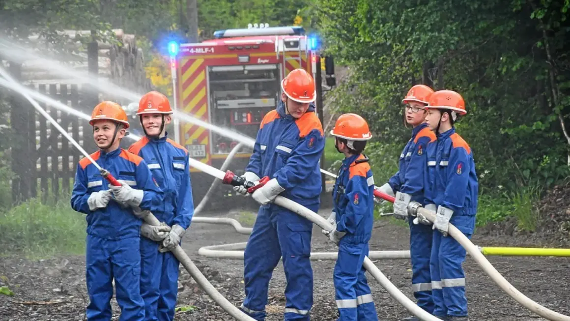 Jugendfeuerwehr übt praktische Ausbildungsideen: Kinder in blauen Uniformen und Helmen halten Wasserschläuche, ein Feuerwehrauto im Hintergrund.