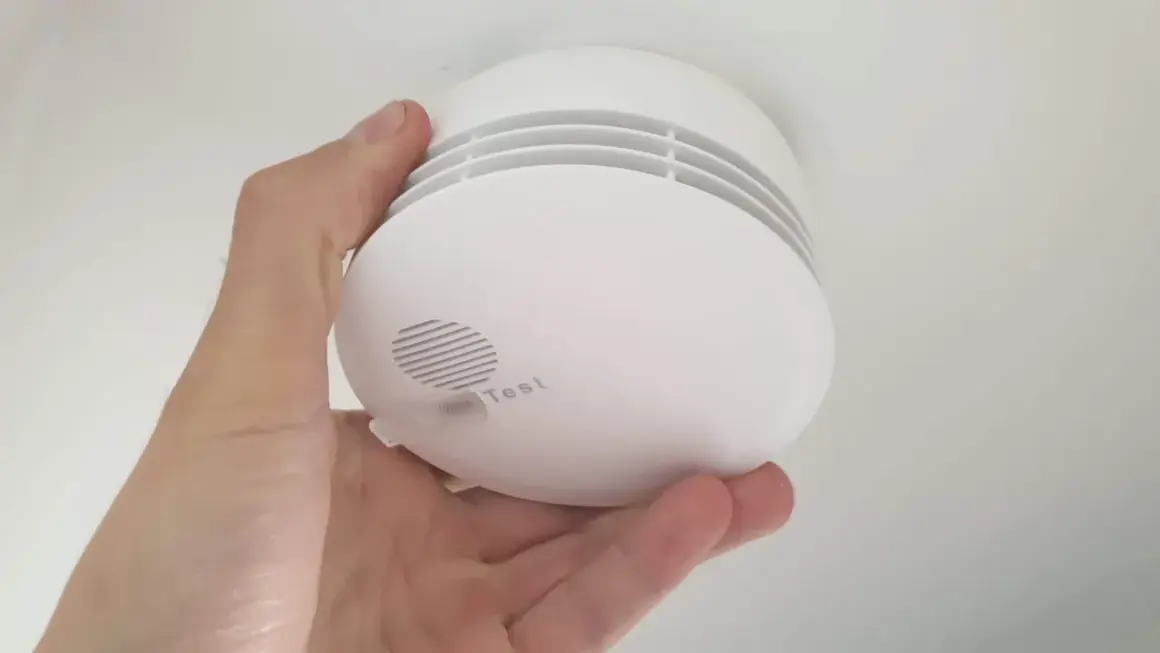 Ein weißer Rauchmelder wird im Keller von einer Hand gehalten. Er hat einen Testknopf und Lüftungsschlitze.