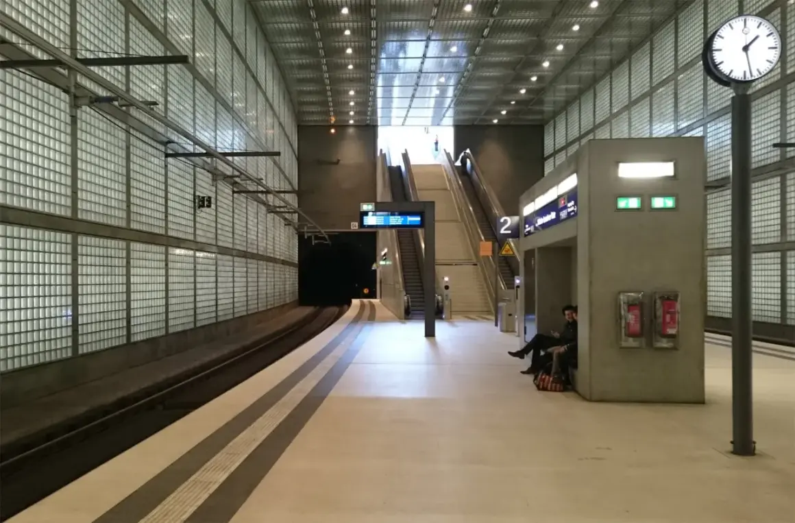 Moderne U-Bahn-Station mit Glaswänden, Rolltreppen und einer Uhr. Die Notbeleuchtung ist gemäß Vorschriften installiert.