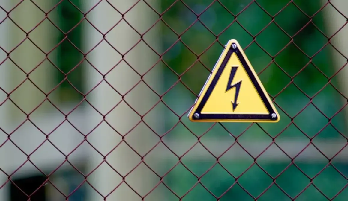 Gefahr! Gelbes Warnschild mit Blitzsymbol auf Drahtzaun. Bei einem leichten Stromschlag was tun? Sofort Abstand halten und Hilfe holen.