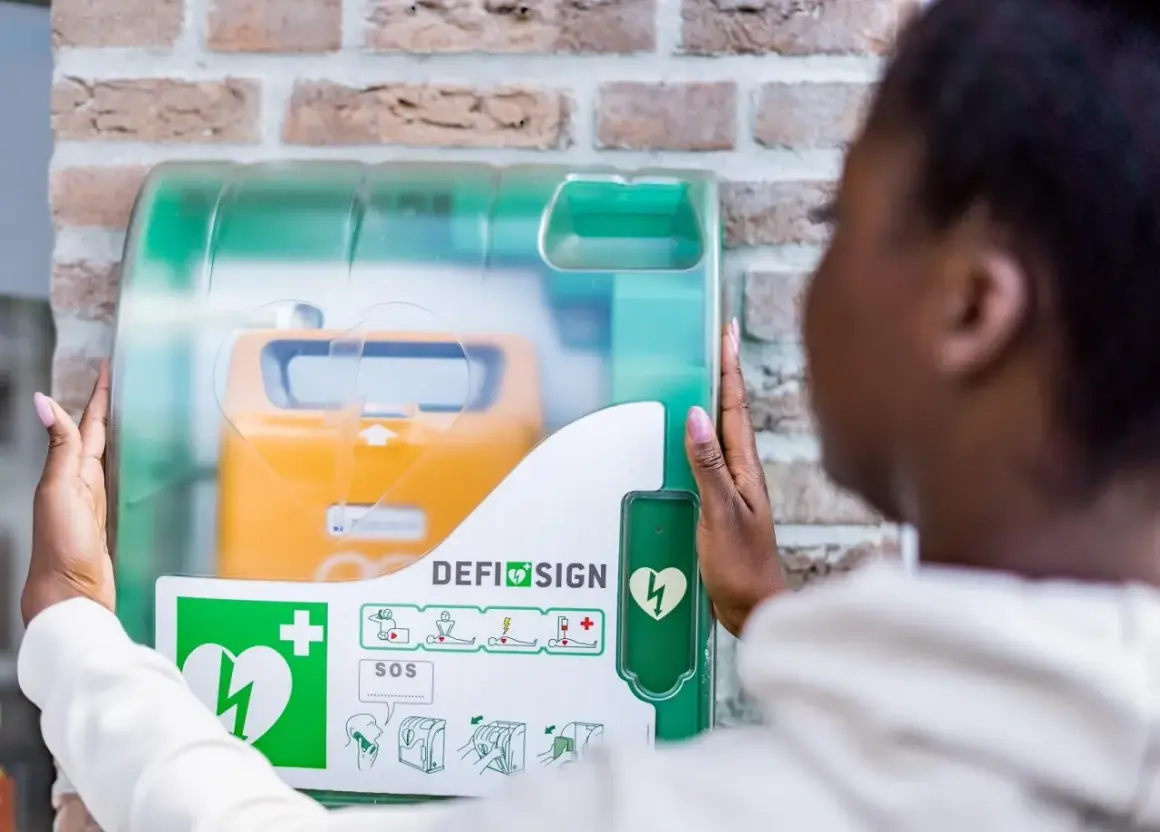 Frau öffnet Defibrillator-Gehäuse, um den Standort zu finden. Gelbes Gerät mit Herzsymbol sichtbar.