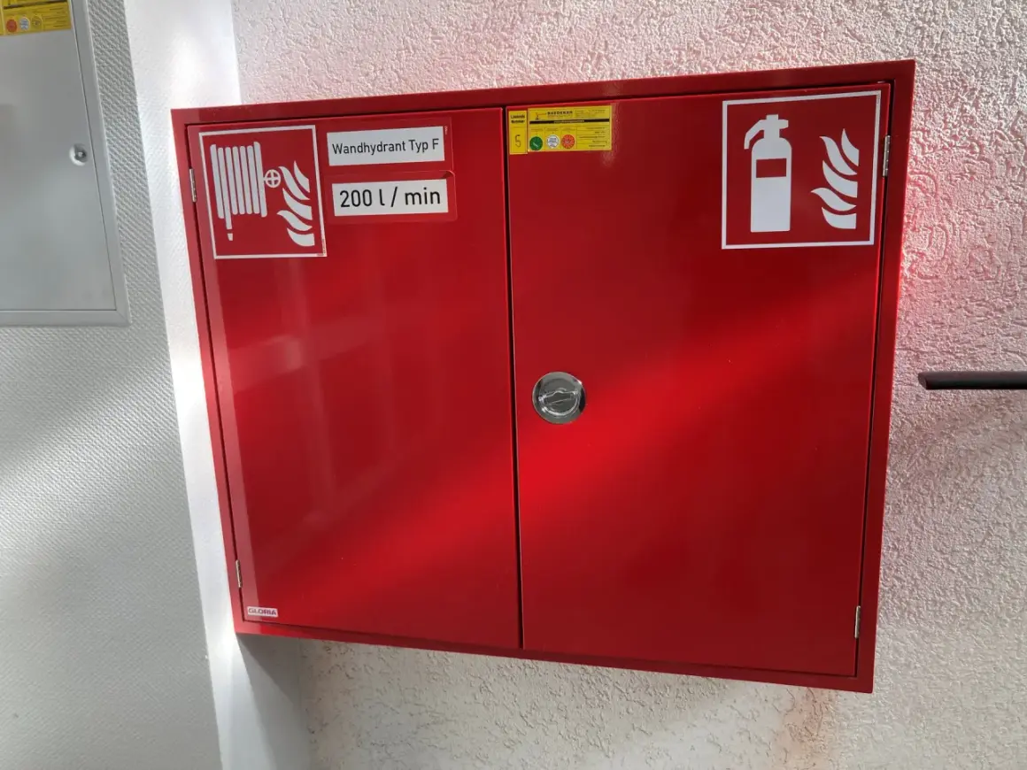 Roter Schrank für Brandschutz in einer Arztpraxis: Wandhydrant Typ F, 200 l/min, und Feuerlöscher-Symbol.