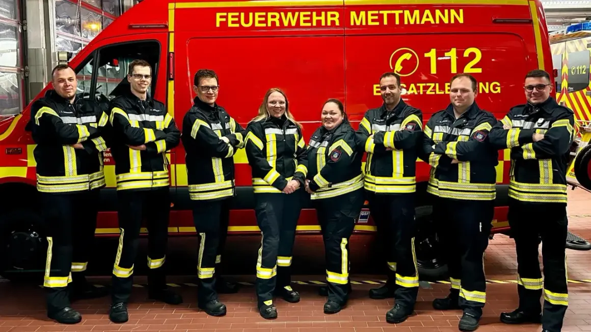 Feuerwehrleute in Mettmann, alle Altersgruppen, stehen stolz vor ihrem roten Einsatzfahrzeug.
