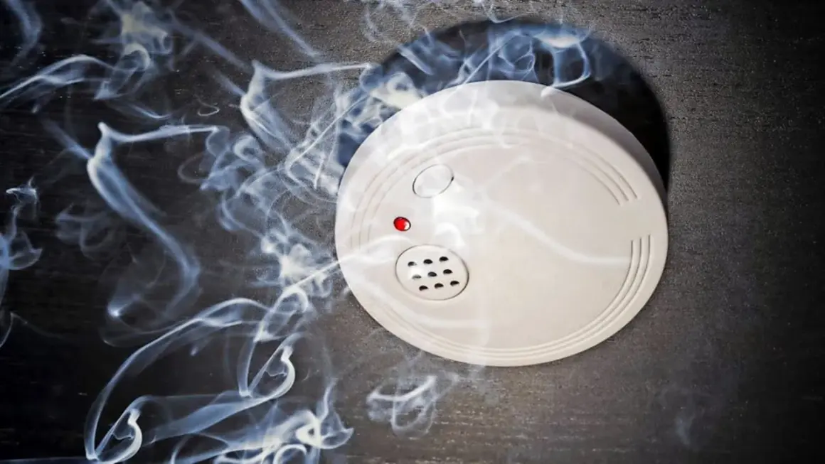 Ein weißer rauchmelder oder feuermelder mit roter LED auf dunklem Grund, umgeben von aufsteigendem Rauch.