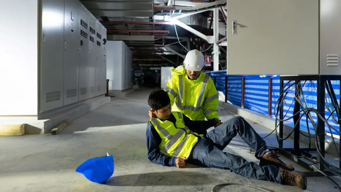 Ein Arbeiter hilft einem Kollegen, der nach einem Stromschlag auf dem Boden liegt. Der helfende Arbeiter trägt eine Warnweste und einen Helm.