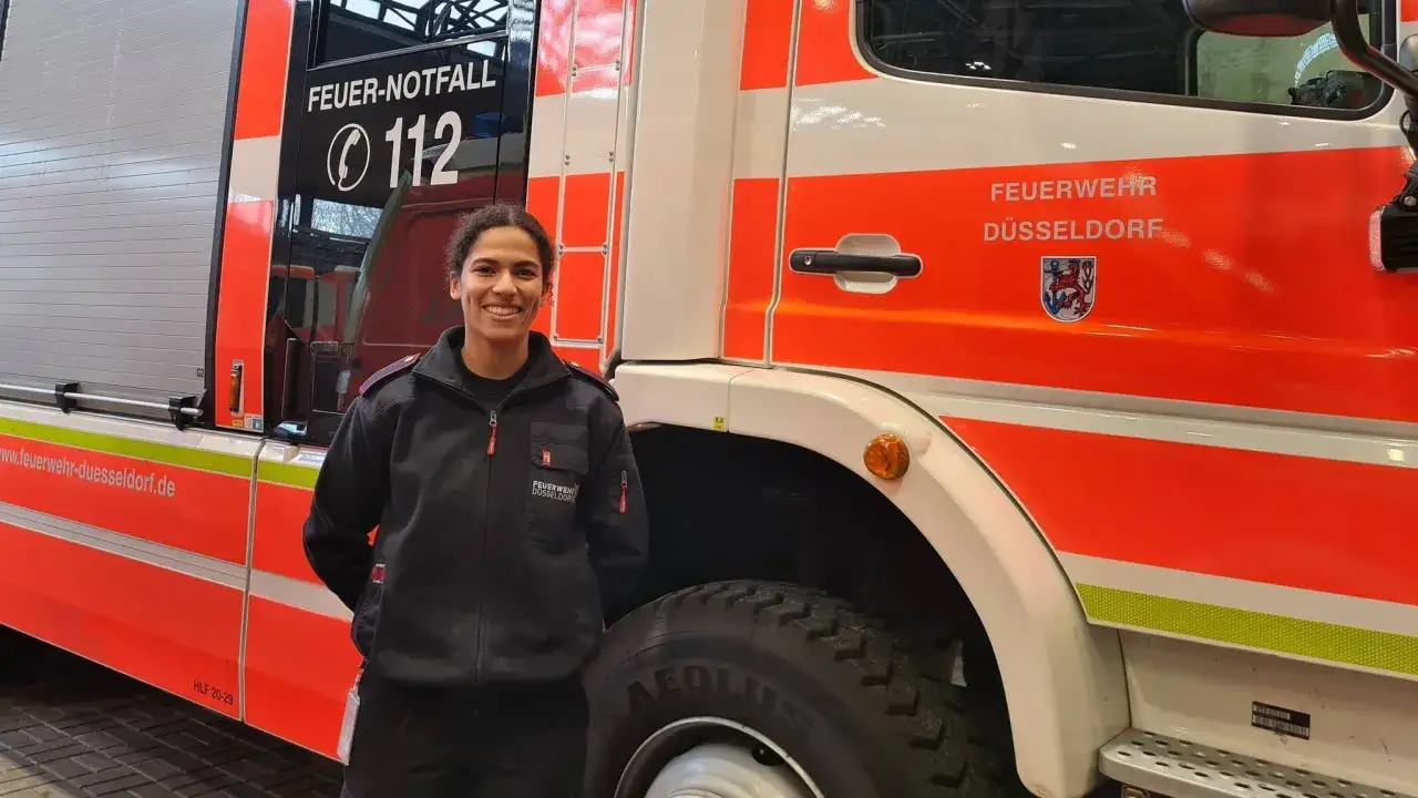Eine junge Frau in Uniform steht vor einem Feuerwehrauto in Düsseldorf. Sie lächelt und wirkt stolz. So wird man Feuerwehrfrau!