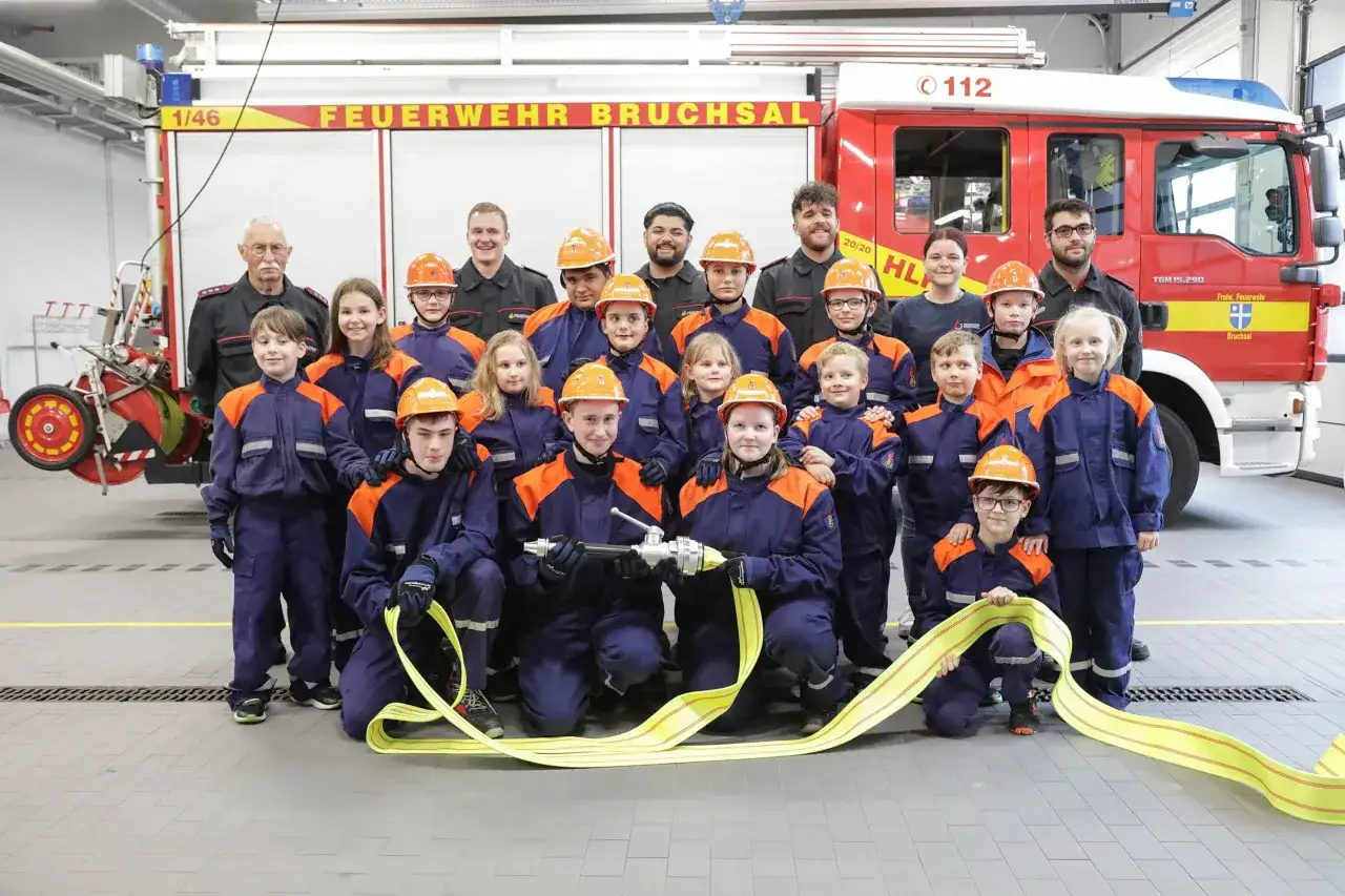 Die Jugendfeuerwehr Bruchsal, mit Kindern und Betreuern, posiert vor einem Feuerwehrauto. Sie tragen Einsatzkleidung und Helme.