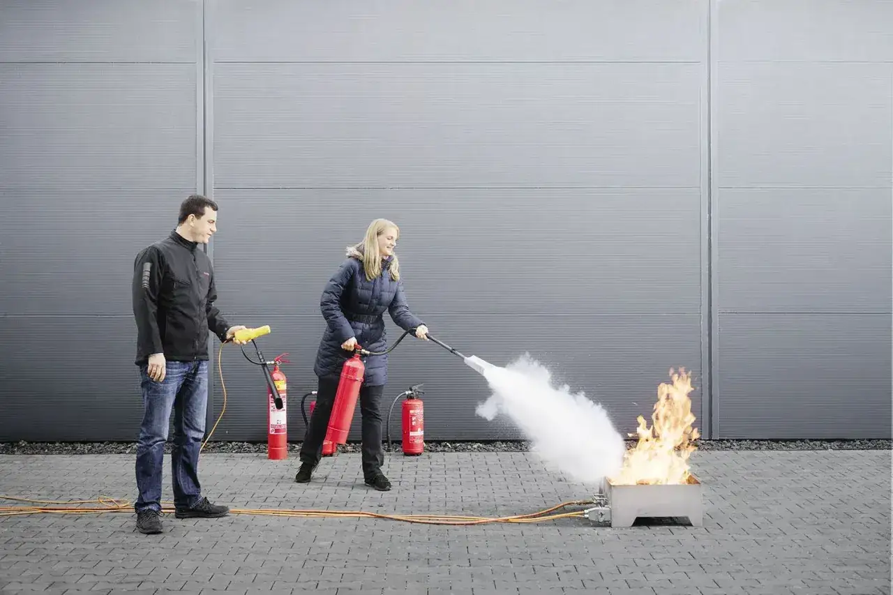 Frau löscht Feuer mit Feuerlöscher, Mann assistiert. Übung für Brandschutzunterweisung Vorlage.