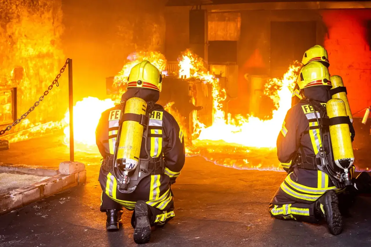 Zwei Feuerwehrleute in Schutzkleidung knien vor einem Inferno. Sie sind bereit, die gefährlichen Berufe bei der Feuerwehr auszuüben und Leben zu retten.