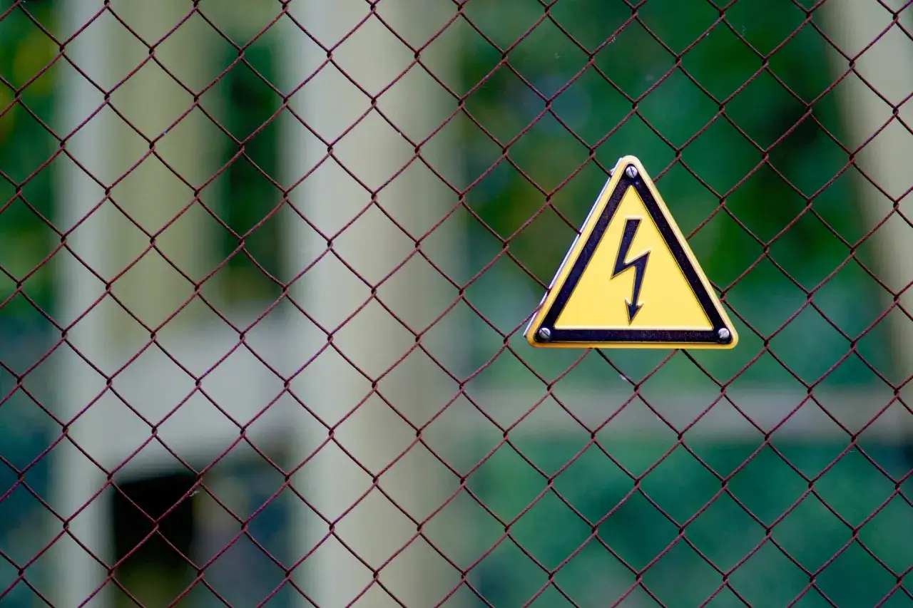 Warnschild mit Blitzsymbol auf Maschendrahtzaun. Achtung vor Stromschlag, Symptome können vielfältig sein.