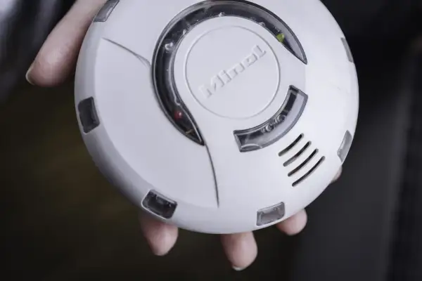 Ein weißer enocean rauchmelder mit dem Schriftzug "Minol" wird von Fingern gehalten. Ein rotes und ein gelbes Licht leuchten.